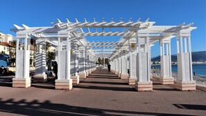 Une pergola blanche s’étend le long d’une promenade côtière, sous un ciel d’un bleu limpide.