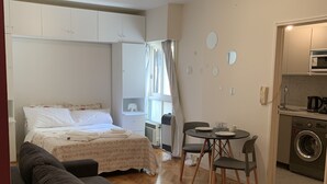 Un dormitorio con cama, sofá y zona de comedor con sillas y mesa. Hay una lavadora en la esquina.