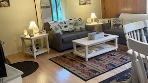 Sala de estar con sofá, mesa de centro y mesitas auxiliares. Cuenta con una ventana con cortinas, un reloj en la pared y una alfombra en el suelo.