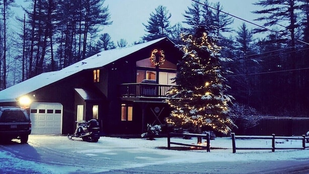 Una cabaña con techo nevado, un árbol de Navidad iluminado, una motocicleta estacionada y un coche en la entrada.