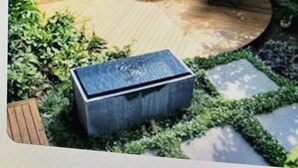 Un patio jardín con dos sillas azules y una mesa pequeña, rodeado de follaje y un sendero de piedra.
