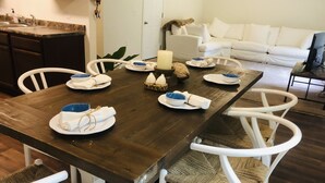 Una mesa de comedor dispuesta con platos, boles y servilletas. Alrededor de la mesa hay sillas blancas con asientos de mimbre. Se ve un sofá blanco y un televisor al fondo.