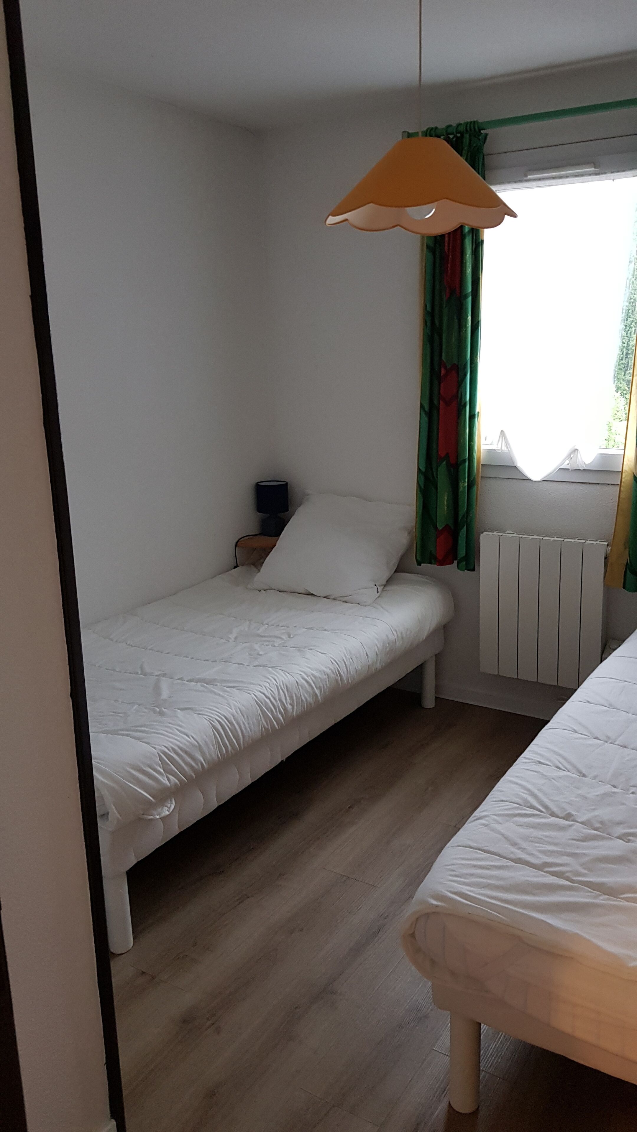 A small, single-bed room with a wooden floor, a bedside table with a lamp, a radiator, and a window with curtains.