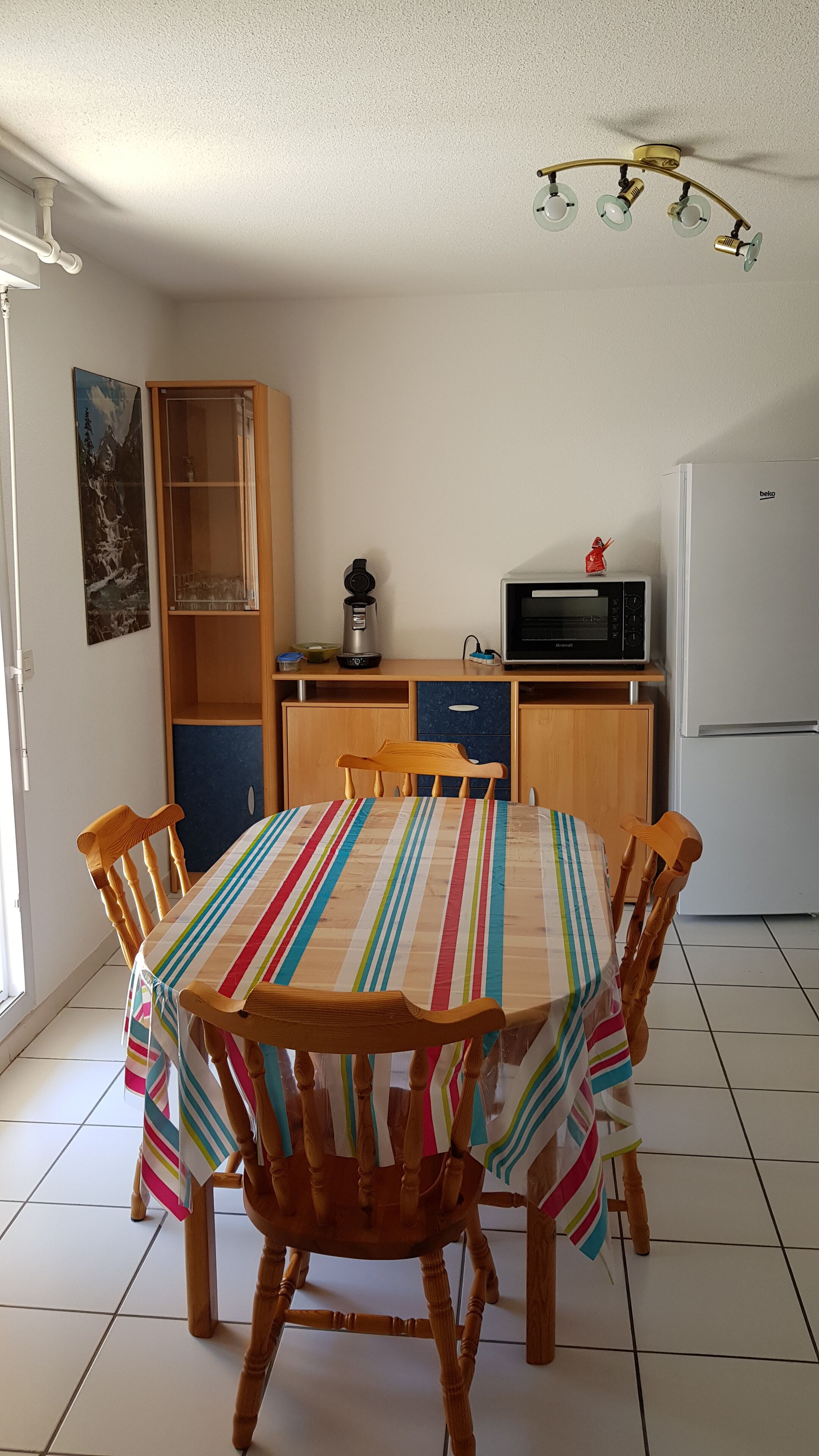 Een eethoek met een strepen tafelkleed, houten stoelen, een koffiezetapparaat, een magnetron en een koelkast.