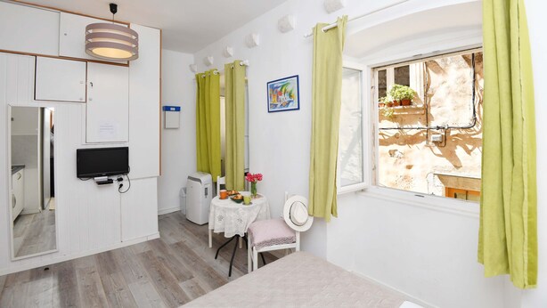A compact room with a dining table, a small white chair, a television mounted on the wall, and a window with green curtains. - Apartments Mila - Studio apartment (Dubrovnik)
