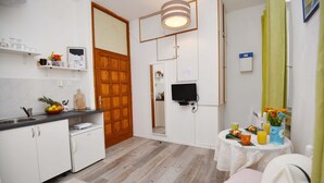 A compact kitchen with white cabinets, a wooden door, a small table with breakfast items, and a TV mounted on the wall. - Apartments Mila - Studio apartment (Dubrovnik)