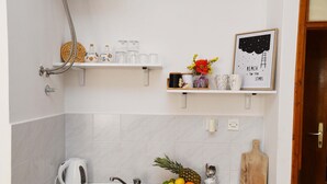 A kitchen with a stainless steel sink, a kettle, a fruit bowl, and a wooden cutting board. There are white shelves with various items and a framed picture on the wall. - Apartments Mila - Studio apartment (Dubrovnik)