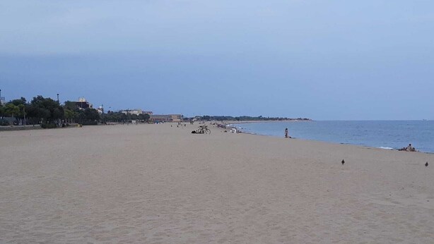 Une vaste plage de sable fin, une mer calme, quelques personnes et des bâtiments en arrière-plan.