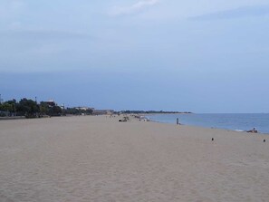 A wide sandy beach with a calm sea, a few people, and buildings in the background.
