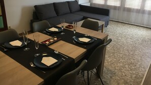 Un coin repas avec une table dressée pour quatre personnes, comprenant des assiettes, des couverts et des verres. On aperçoit un canapé au fond et une fenêtre avec des stores.