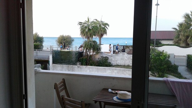 A balcony with a wooden chair and table, overlooking a beach with palm trees and people. - House 20 meters from the sea! (Fossacesia Marina)