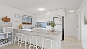 Une cuisine moderne avec des meubles blancs, un grand îlot central doté de tabourets, une horloge et une fenêtre donnant sur une maison.