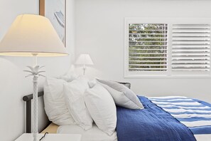A bedroom with a bed, pillows, a nightstand, a lamp, and a window with blinds.