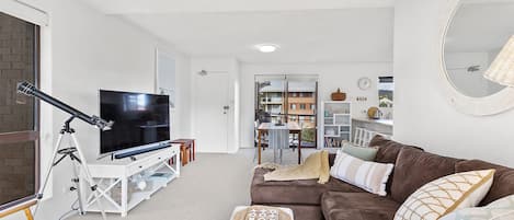 Una sala de estar moderna con un televisor de pantalla plana, un sofá marrón, un reposapiés blanco y un área de comedor al fondo.
