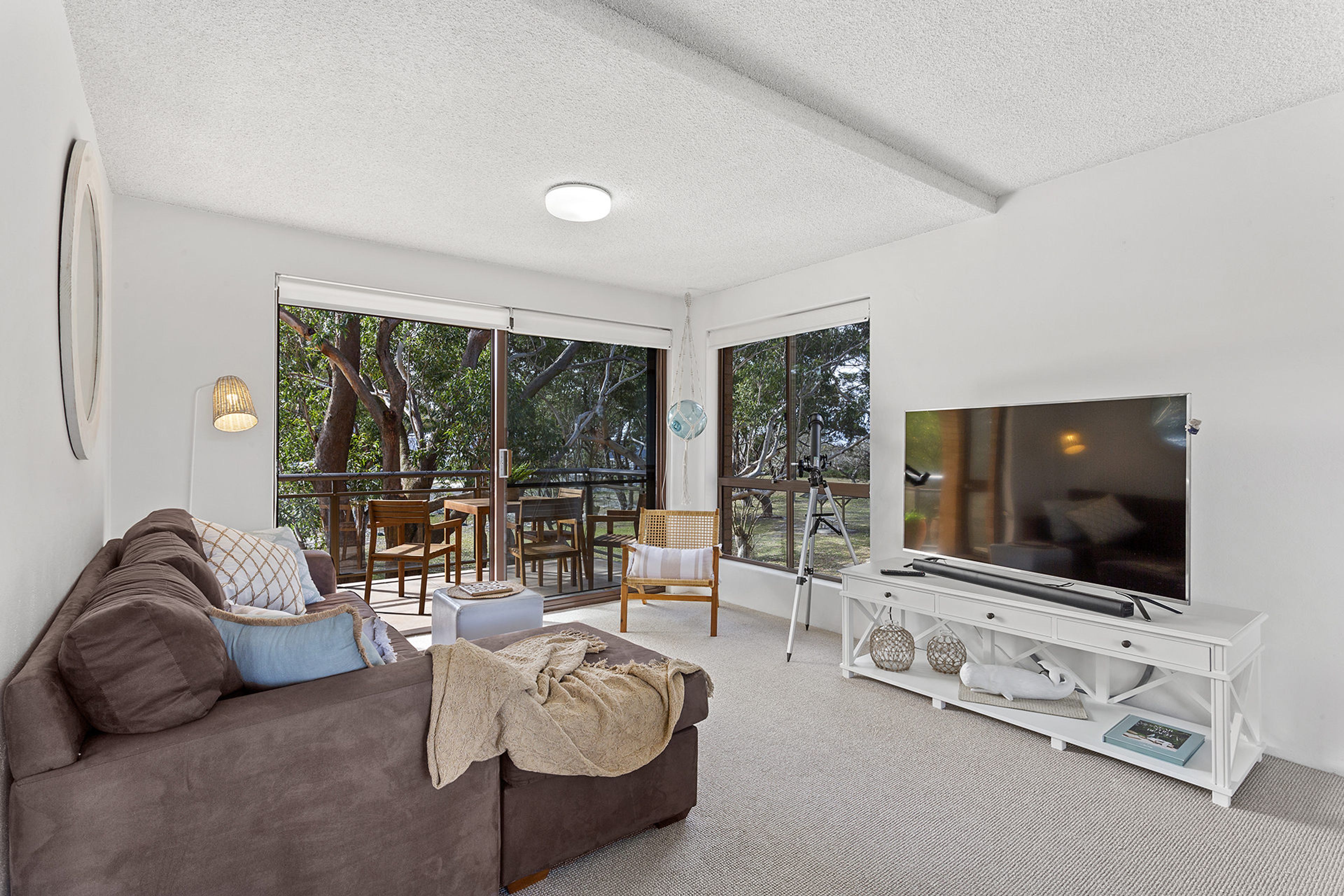A spacious living room with a large flat-screen TV, a brown sofa, and a balcony with outdoor furniture.