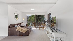 Un salon moderne avec un canapé marron, une table basse blanche et un téléviseur posé sur un meuble blanc. Une grande fenêtre offre une vue sur les arbres, et un balcon est également visible.