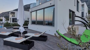 Zona relax esterna moderna con lettini e ombrelloni, ampie finestre e montagne sullo sfondo.
