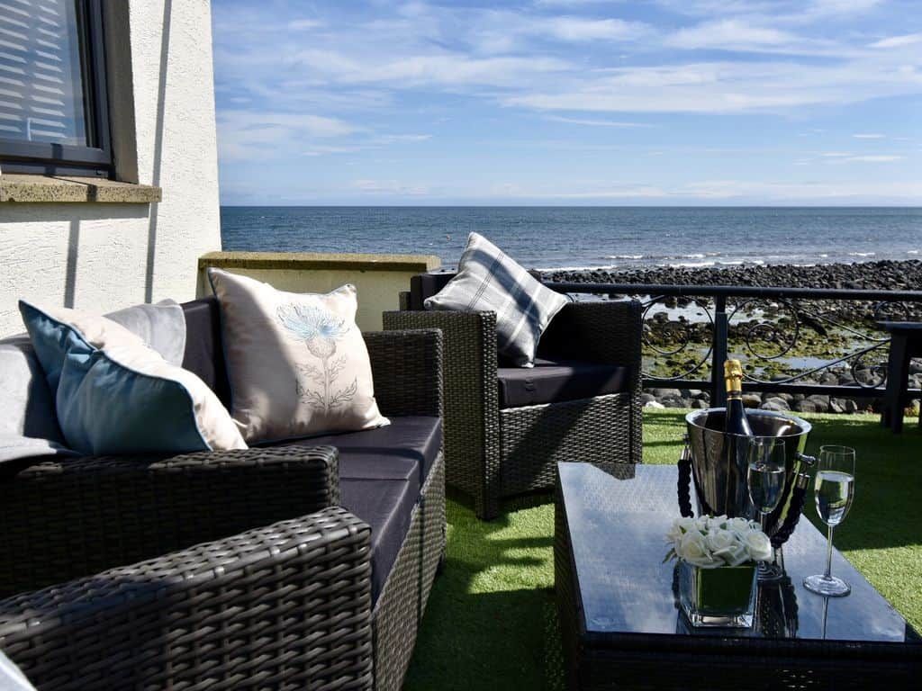 A seaside patio with wicker furniture and a table set with a bottle, glasses, and flowers.