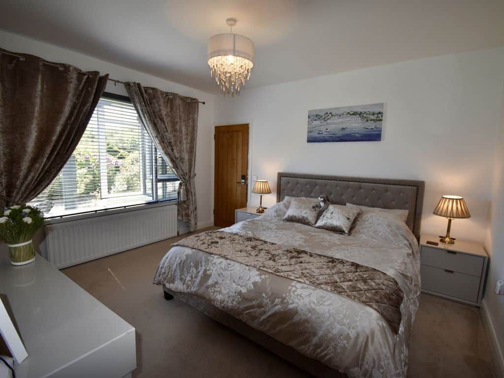 A bedroom with a large bed, two bedside lamps, a window with curtains, a painting on the wall, and a radiator.