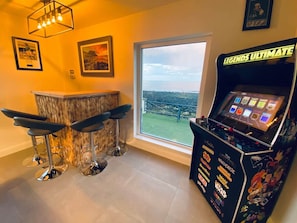 Zona de bar con tres taburetes, una barra y una máquina de juegos con la etiqueta 'LEGENDS ULTIMATE'.