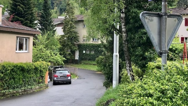 Une rue résidentielle où une voiture est garée sur le bord, entourée d’une végétation luxuriante et de maisons.