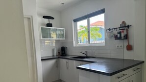Una cocina moderna con armarios blancos, una encimera negra, una ventana con vistas a palmeras y un estante pequeño con utensilios de cocina.