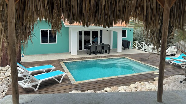 Zona de piscina con sillones, terraza de madera y edificio con techo de paja.