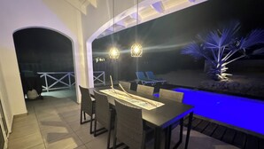 Un espacio de comedor al aire libre con piscina, dos lámparas colgantes y vistas nocturnas de la piscina.