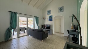 Un salón amplio con un sofá gris, un televisor y una mesa de comedor. Hay una puerta corrediza de vidrio que da a un patio exterior.