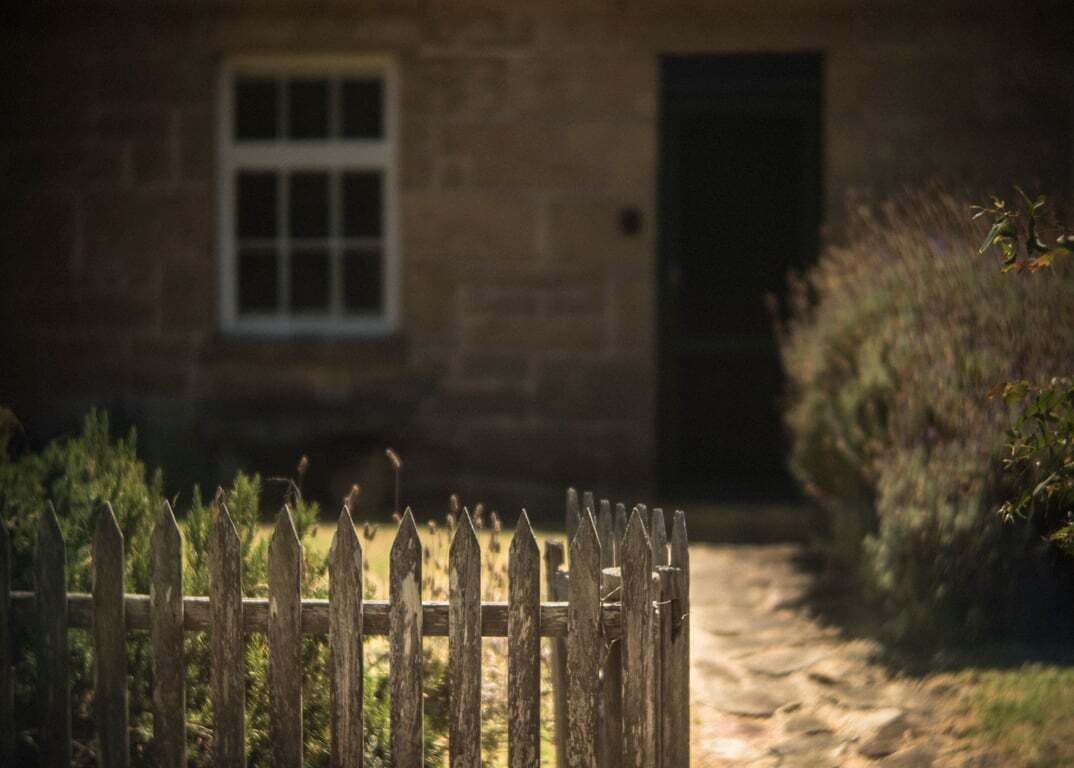 A stone building with a window and a door, surrounded by a wooden fence and vegetation.