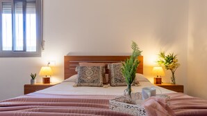 Ein ordentlich bezogenes Bett mit einer rosa Decke, zwei Nachttischlampen, einer Vase mit einer Pflanze und einem kleinen Tisch mit einer Tasse.