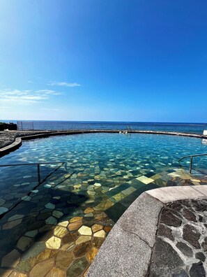 A clear pool with a stone pattern bottom, a metal railing, and a view of the sea.