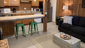 A compact kitchen with wooden cabinets, a microwave, a toaster, and a refrigerator. There are two green stools and a black sofa with a cushion. A small table with a tray and a remote control is on the floor.