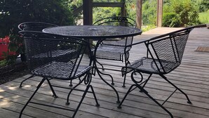 A covered outdoor patio with a round metal table and four chairs, surrounded by wooden decking and lush greenery.