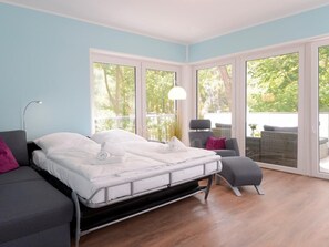 A modern bedroom with a bed, a sofa, and a chair. There are large windows and a balcony with outdoor furniture. - Tide App. 4 dike light (Ostseebad Boltenhagen)