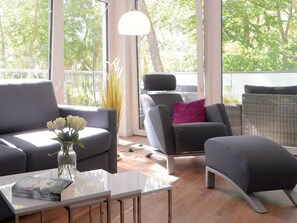 Ein modernes Wohnzimmer mit Sofa, Sessel, Hocker und Couchtisch. Auf dem Couchtisch steht eine Vase mit Blumen.