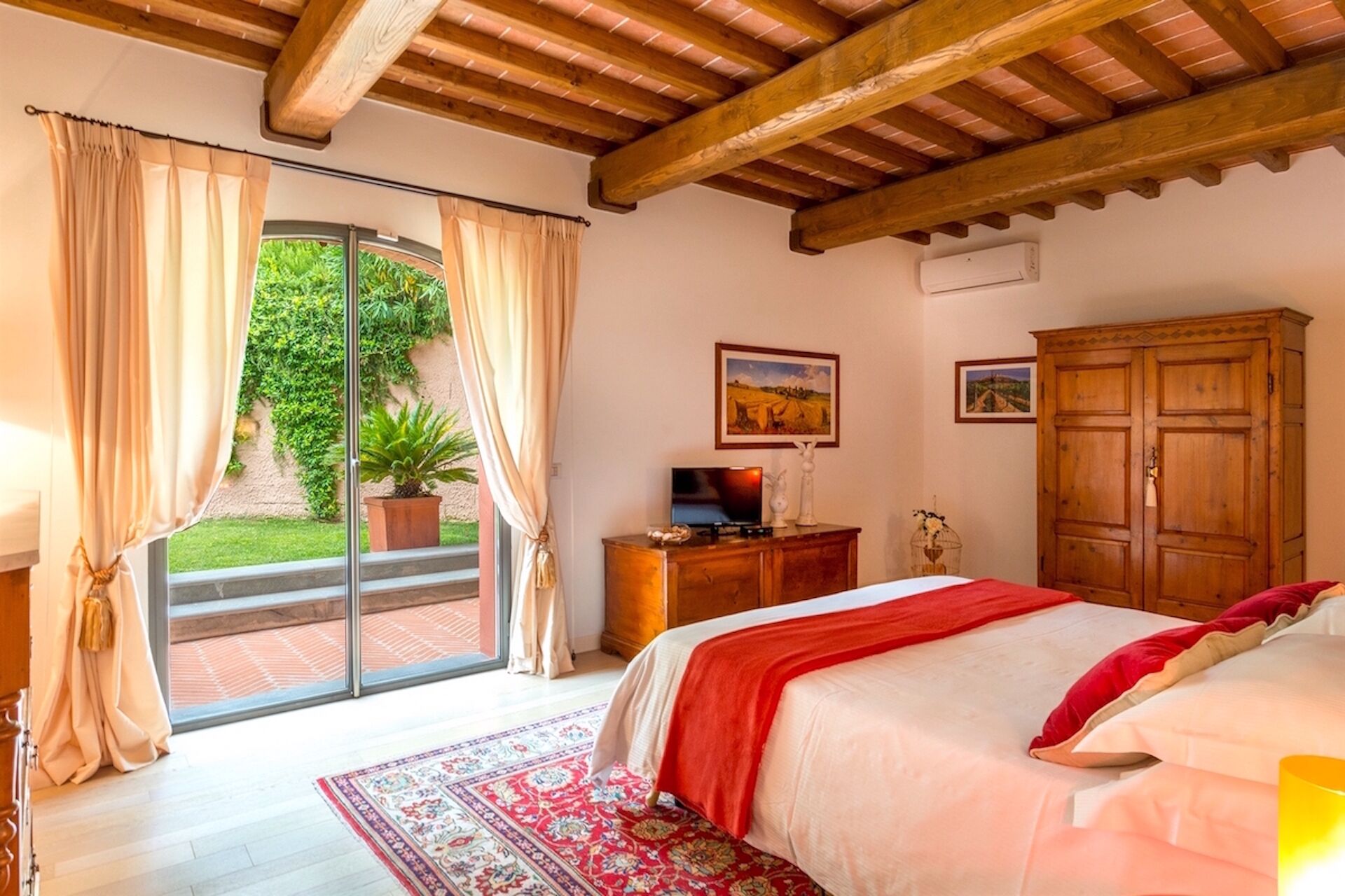 Un dormitorio con una cama, un armario de madera, un televisor sobre un mueble de madera y un ventanal corredero que da a un jardín.