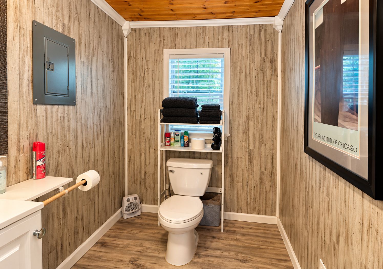 A bathroom with wooden paneling, a toilet, a shelf with towels, and a framed picture on the wall.
