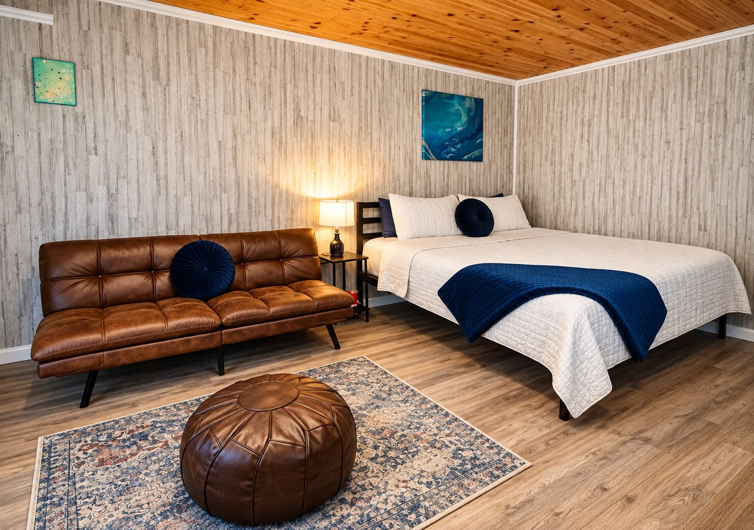 A bedroom with a brown leather sofa, a bed with a blue blanket, a wooden floor, and a wooden ceiling.