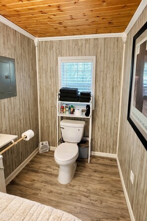A compact bathroom with a toilet, a window, and a shelf with toiletries. - The Cozy King Studio #2 (Benton)