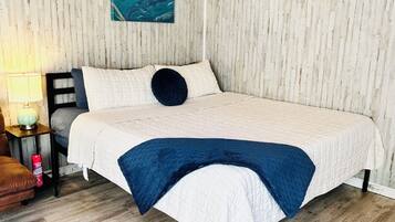 Un dormitorio con techo de madera, cama con colcha blanca y azul, almohada oscura, mesita de noche con lámpara y alfombra en el suelo.