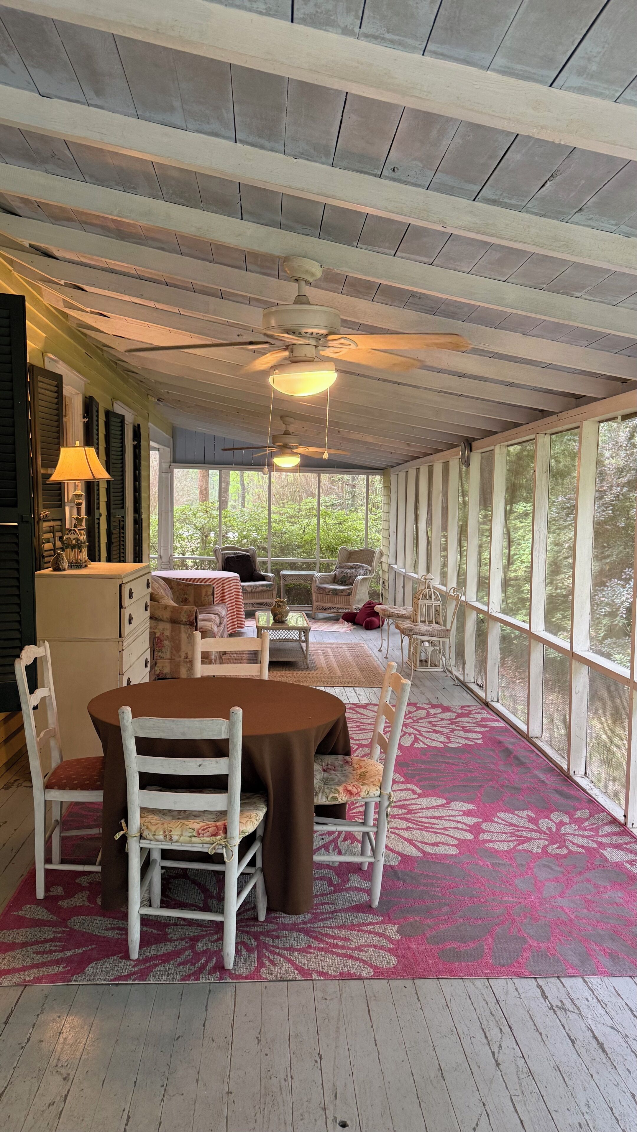 Une véranda abritée avec une table et des chaises, un ventilateur de plafond et une vue sur la verdure environnante.
