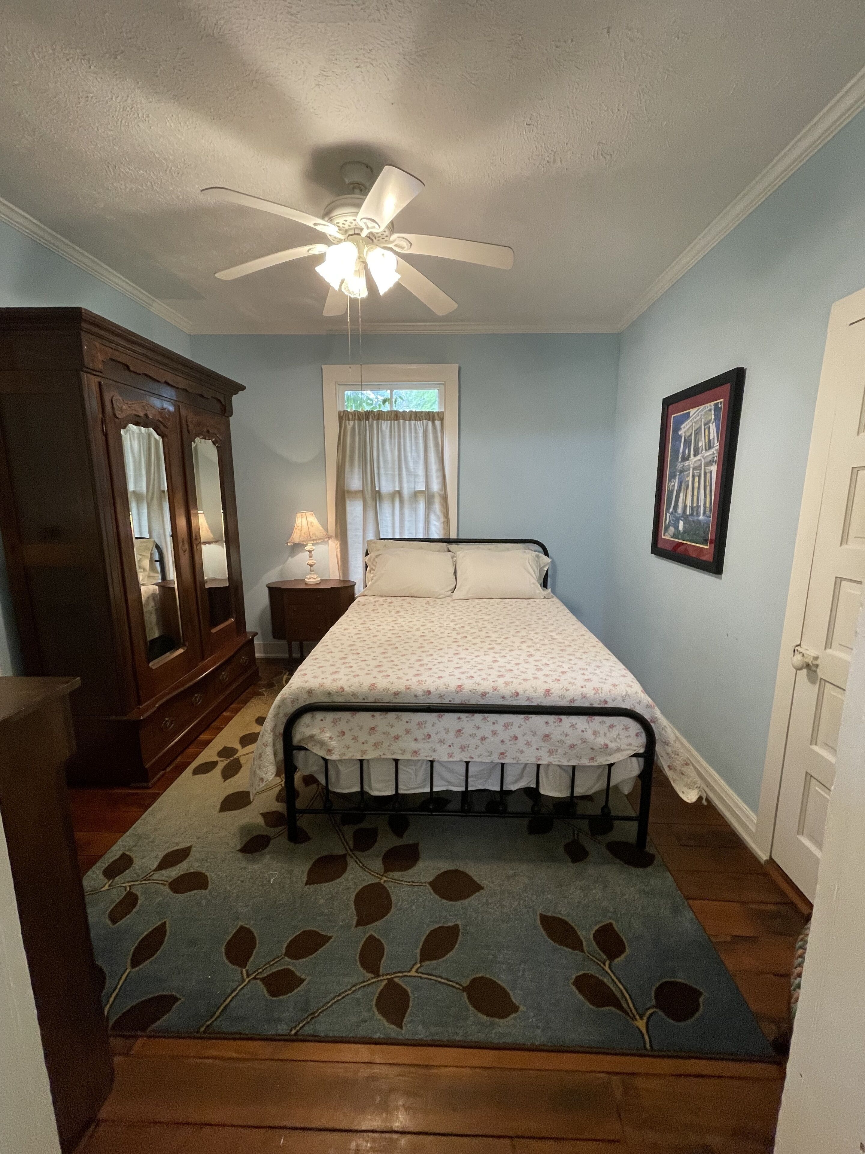Une chambre à coucher avec un lit, un ventilateur de plafond, une fenêtre avec des rideaux, une commode, un tableau encadré au mur et une porte.