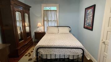 Une chambre à coucher avec un lit, un ventilateur de plafond, une fenêtre avec des rideaux, une commode, un tableau encadré au mur et une porte.