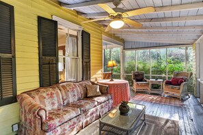 A porch with a floral sofa, wicker chairs, a table with a vase, and a ceiling fan.