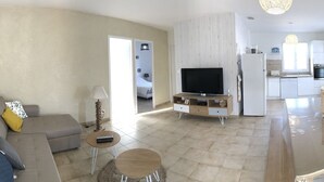 A modern living room with a sofa, coffee table, and TV. A kitchen is visible in the background with a refrigerator and cabinets. - Renovated house, 300m from the beach, with a boule court (La Brée-les-Bains)