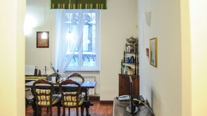 Una habitación con piso de madera, una mesa de comedor con sillas, un ventanal con cortina verde y una estantería.