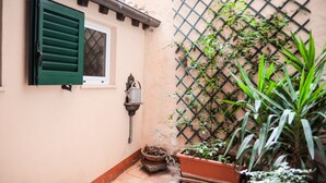 Un patio estrecho con una ventana y contraventanas verdes, un aplique de pared y plantas en macetas.
