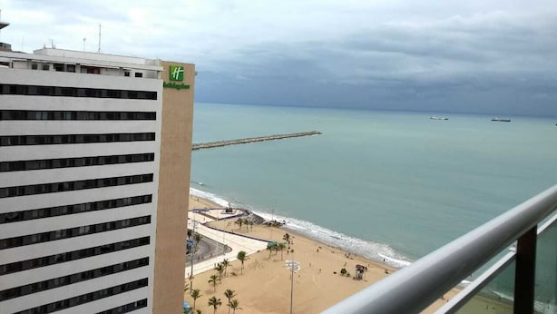 Vista de una playa con un muelle que se adentra en el mar, arena y un edificio con balcón.