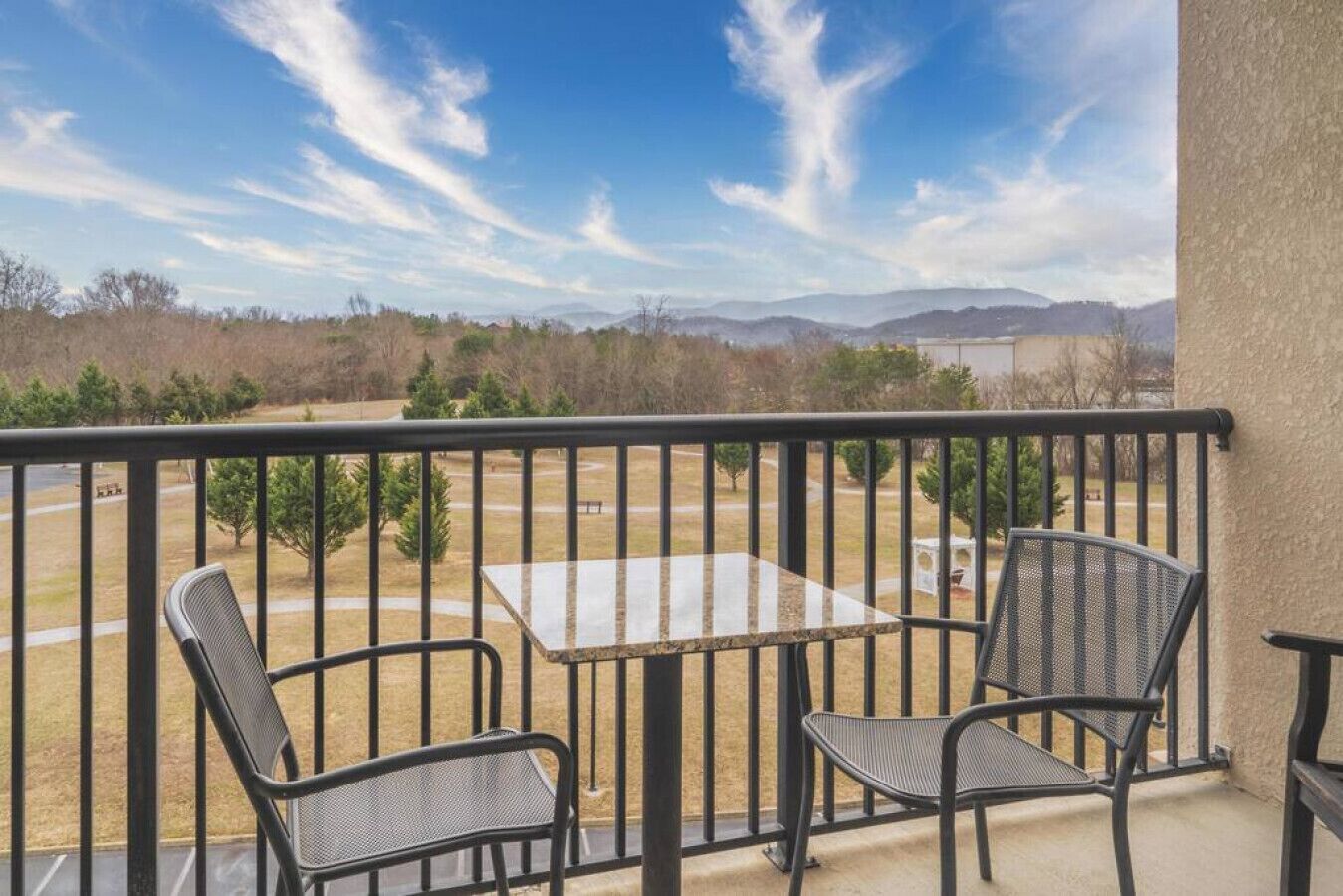A balcony with a table and two chairs, overlooking a grassy area with trees and distant mountains.